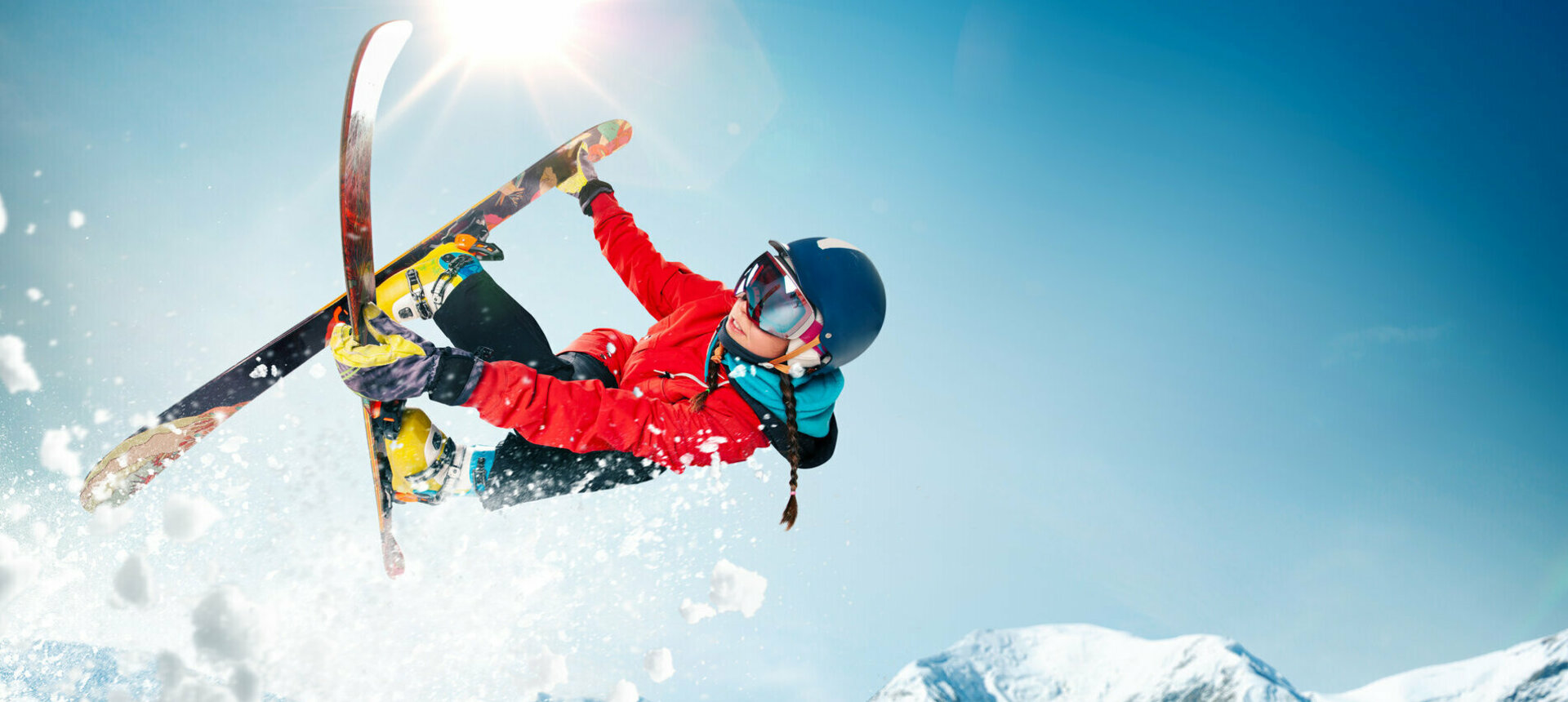 Weibliche Skifahrerin mit Tricksprung, dahinter Sonne und verschneite Berge
