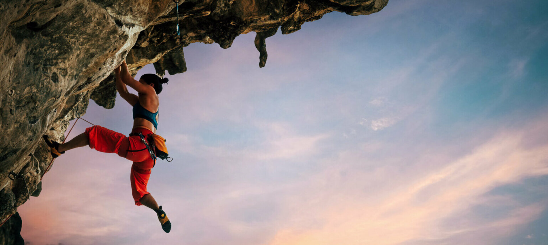 Weibliche Kletterin klettert an Überhang, dahinter Sonnenuntergang